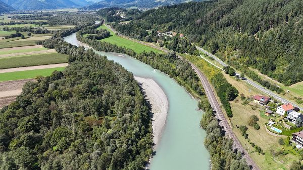 Luftaufnahme der Drau vor ihrer Revitalisierung in einem alpinen Tal. Der Fluss verläuft in einem breiten, vergleichsweise gleichförmigen Bett durch eine von Landwirtschaft geprägte Landschaft mit Feldern, Wiesen und bewaldeten Uferstreifen. Entlang des rechten Ufers verlaufen eine Bahnstrecke und eine Straße mit einzelnen Wohnhäusern. Bewaldete Berghänge und hohe Gipfel prägen die Kulisse unter einem teilweise bewölkten Himmel.