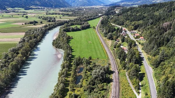 Luftaufnahme der Drau vor ihrer Revitalisierung in einem alpinen Tal. Der Fluss verläuft in einem breiten, vergleichsweise gleichförmigen Bett durch eine von Landwirtschaft geprägte Landschaft mit Feldern, Wiesen und bewaldeten Uferstreifen. Entlang des rechten Ufers verlaufen eine Bahnstrecke und eine Straße mit einzelnen Wohnhäusern. Bewaldete Berghänge und hohe Gipfel prägen die Kulisse unter einem teilweise bewölkten Himmel.
