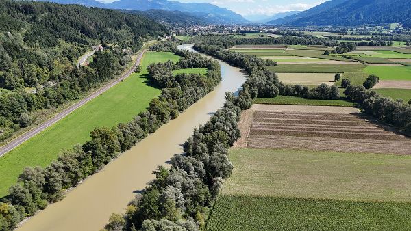 Luftaufnahme der Drau vor ihrer Revitalisierung in einem alpinen Tal. Der Fluss verläuft in einem breiten, vergleichsweise gleichförmigen Bett durch eine von Landwirtschaft geprägte Landschaft mit Feldern, Wiesen und bewaldeten Uferstreifen. Entlang des linken Ufers verlaufen eine Bahnstrecke und eine Straße mit einzelnen Wohnhäusern. Bewaldete Berghänge und hohe Gipfel prägen die Kulisse unter einem teilweise bewölkten Himmel.