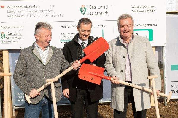 drei Herren vor einer Bautafel, die außenstehenden halten einen Spaten schraeg zur Mitte 