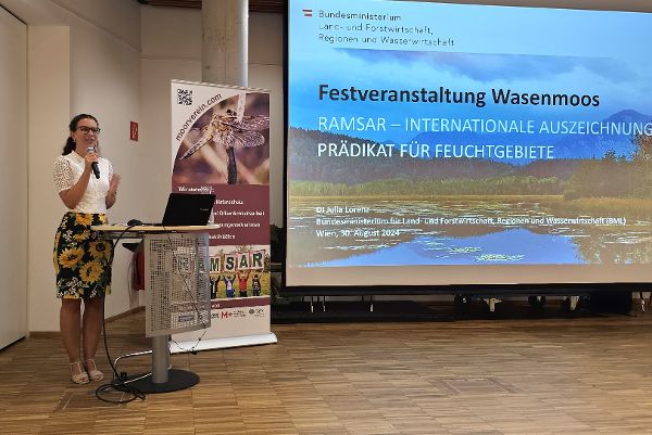 links am Rednerpult Dipl.Ing. Julia Lorenz (BML) und rechts eine große Leinwand mit Foto Wasenmoos und Beschriftung