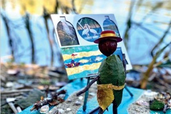 Eine aus Naturmaterialien gebastelte Figur eines Künstlers steht auf eine Leinwand malend am Rande einer Wasserfläche