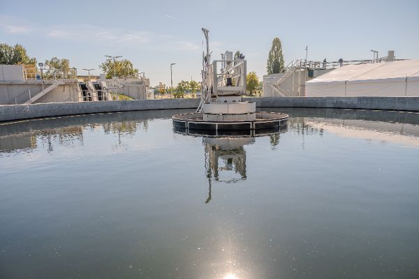 Blick auf das runde Nachklärbecken der Kläranlage Podersdorf anlässlich der Eröffnung. In der Mitte des Beckens befindet sich die mechanische Räumeranlage, deren Konstruktion sich im Wasser spiegelt. Im Hintergrund sind weitere Anlagenteile, Betriebsstege und ein Festzelt zu sehen.