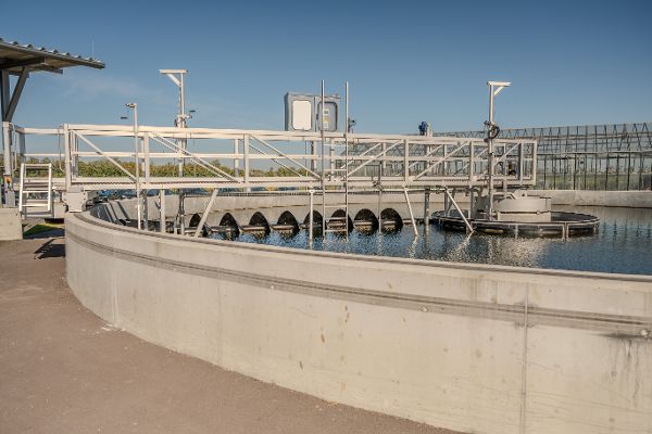 Ein großes, kreisrundes Nachklärbecken aus hellem Beton in der neuen Kläranlage Podersdorf am See unter blauem Himmel. Über das wassergefüllte Becken erstreckt sich eine begehbare Metallbrücke mit Geländer. Im Becken befinden sich mechanische Vorrichtungen zur Abwasserreinigung, während im Hintergrund weitere Industrieanlagen zu sehen sind.