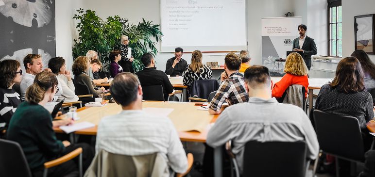 Teilnehmerinnen eines Expertinnenworkshops sitzen in einem Raum, Vortragender steht vor einer Präsentation an der Wand