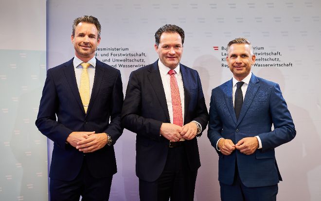 Drei Männer in Anzügen stehen nebeneinander vor einer Wand mit Text und Logos des Bundesministeriums für Landwirtschaft und Forstwirtschaft.