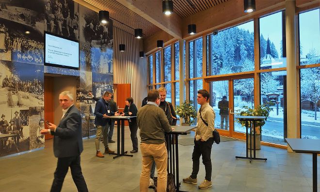 Empfangsszene des Policy Labs in Werfenweng. Leute stehen um Tische und unterhalten sich, auf einem Bildschirm steht "Willkommen zum Policy Lab" und durch das Fenster sieht man die winterliche Kulisse des Ortes Werfenweng.
