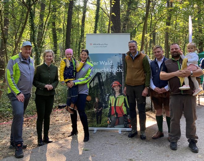 Mehrere Personen stehen vor einem Plakat, auf dem eine Forstarbeiterin abgebildet ist. Im Hintergrund sind Bäume zu sehen.