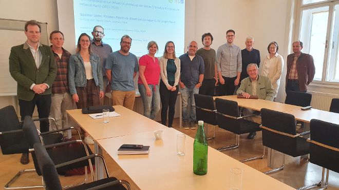 nebeneinander gereihte Menschen in einem in U-Form angneordneten Besprechungstischen, im Hintergrund eine Praesentationsabbildung dieser Arbeitsgruppe