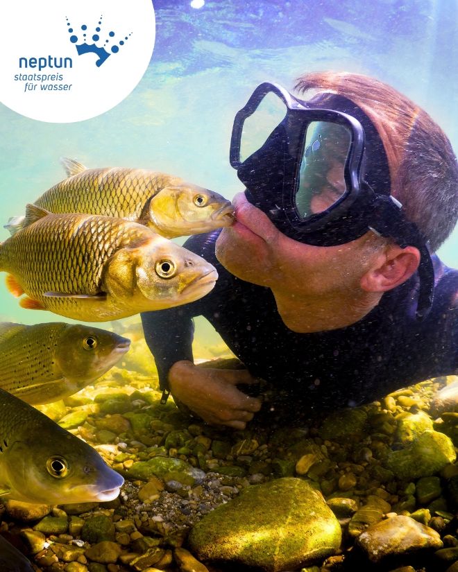 Das Hauptsujet des Neptun Staatspreis 2027 zeigt einen Mann mit Taucherbrille unter Wasser, umgeben von Fischen. Ein Fisch berührt ihn dabei sanft mit einem kleinen Kuss.